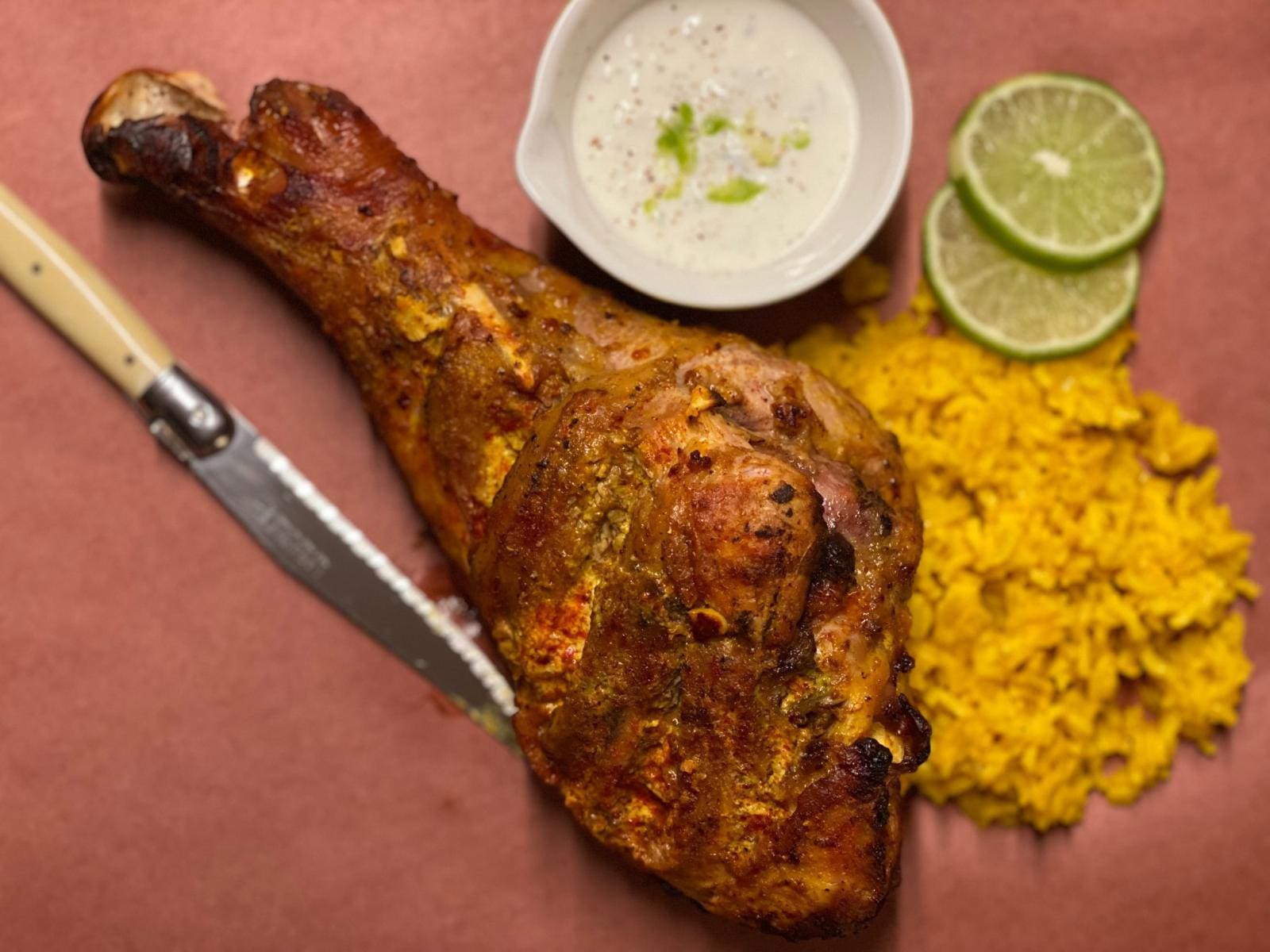 Certified organic, pasture raised turkey drumstick shown roasted on a serving paper with a carving knife alongside turmeric rice, lime slices, and yogurt sauce.