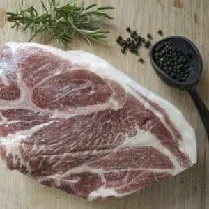 A pasture raised pork Boston butt shoulder roast on a wood cutting board with rosemary and black pepper garnishes.