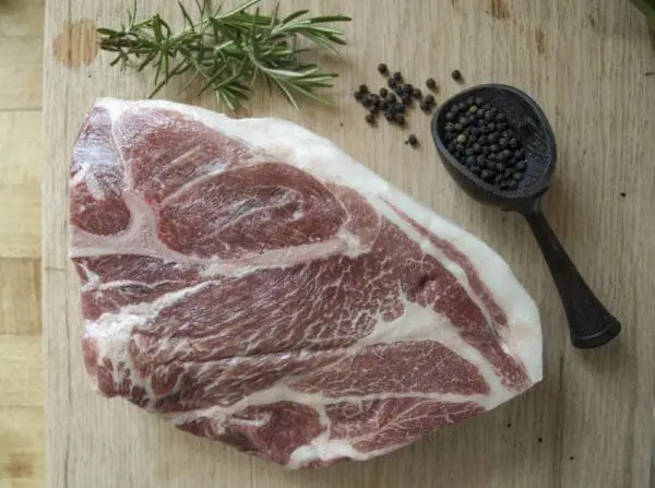 A pasture raised pork Boston butt shoulder roast on a wood cutting board with rosemary and black pepper garnishes.