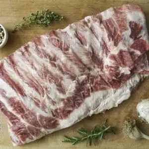 Pasture raised pork spare ribs on a wood cutting board with garnishes and garlic.