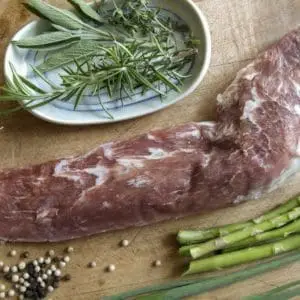 Pasture raised pork tenderloin on a wood cutting board with garnishes and asparagus.