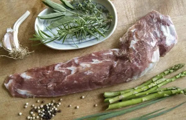 Pasture raised pork tenderloin on a wood cutting board with garnishes and asparagus.