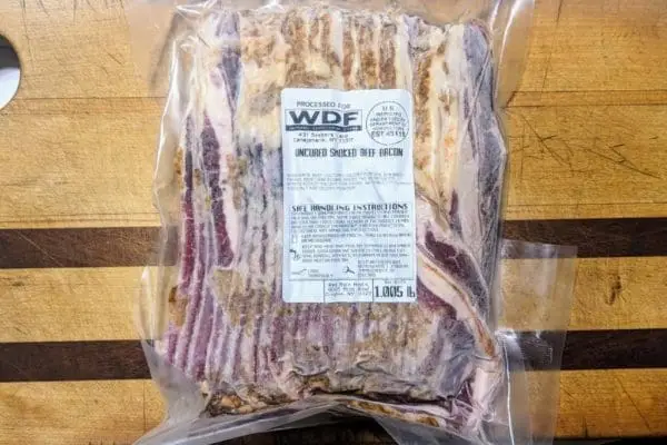 A view of a one pound package of sliced grass fed beef bacon on a wood cutting board.